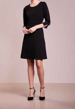 Vrouw draagt een knielange zwarte jurk met textuur, driekwart mouwen en zwarte hoge hakken met enkelriempje, staande tegen een effen achtergrond.