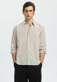 Camicia a maniche lunghe beige chiaro con sottili righe verticali, chiusura a bottoni e polsini arrotolati, abbinata a pantaloni neri. Tessuto morbido.