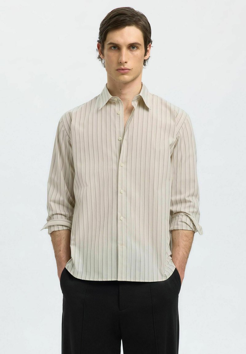 Camicia a maniche lunghe beige chiaro con sottili righe verticali, chiusura a bottoni e polsini arrotolati, abbinata a pantaloni neri. Tessuto morbido.