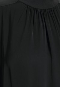 Zblízka na černou látku šatů se nařasenými záhyby pod žebrovaným výstřihem.