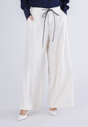 Pantalones blancos de pernera ancha con rayas finas y cordón negro, usados con tacones blancos y top de manga larga azul marino, manos en los bolsillos.