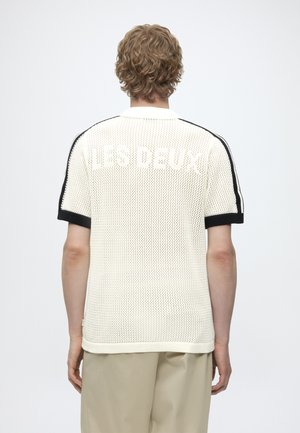 Personne blonde debout, dos à la caméra, portant une chemise blanche en maille à manches courtes avec bordure noire et le texte « LES DEUX » dans le dos.
