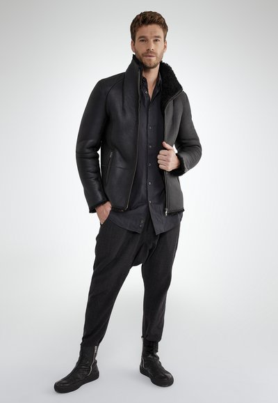 Uomo che indossa un giubbotto in shearling nero, una camicia scura, pantaloni larghi scuri e stivali neri, in piedi con sicurezza contro uno sfondo chiaro e semplice.