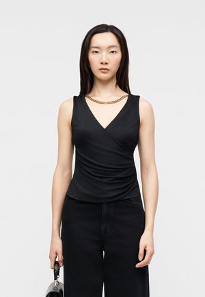 Femme aux longs cheveux noirs portant un haut noir sans manches, un pantalon noir, un collier en chaîne dorée, tenant un sac à main noir sur un fond uni.