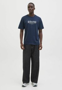 Marineblå bomulds T-shirt med hvid tekst, parret med løstsiddende mørk denim jeans. Modellen bærer grå og hvide træningssko. Enkel, afslappet stil.
