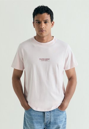 Joven con el pelo corto y rizado que lleva una camiseta rosa claro con el texto "Scalpers Company" y vaqueros azules, manos en los bolsillos, fondo neutro.