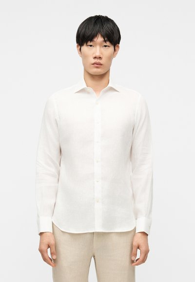 Jeune homme debout, faisant face à l'avant, portant une chemise blanche à manches longues boutonnée et un pantalon beige, sur fond uni.