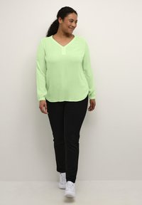 Světle zelená blůza s dlouhým rukávem a výstřihem do V, s volným střihem a elastickými manžetami, spárovaná s černými slim-fit kalhotami a bílými teniskami.
