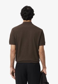 Polo shirt marrone con maniche corte e colletto a coste, visto da dietro. Il tessuto appare morbido e texture, abbinato a pantaloni neri.