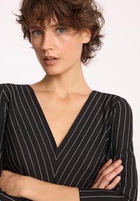 Czarna bluzka z dekoltem w serek w białe pionowe paski, z długimi rękawami i dopasowanym krojem. Modelka ma skrzyżowane ramiona i krótkie włosy.