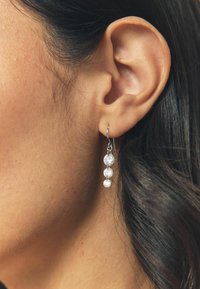 Kõrvalvaade kõrvale, kus on hõbedast ripats-kõrvarõngas kolme ümmarguse läbipaistva kalliskiviga, tumeda juukse ja loomuliku nahatooniga taustal.