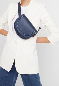 Granatowa skórzana torba na ramię z półokrągłą klapką, fakturowaną powierzchnią i regulowanym paskiem, noszona na białej bluzce i marynarce.
