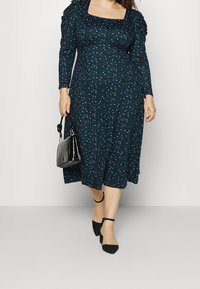 Robe à manches longues, vert foncé avec un petit imprimé floral coloré, encolure carrée, corsage ajusté, jupe fluide. Associée à un sac à main noir et des chaussures noires.