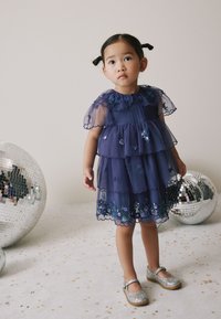 Vestido azul marino de varios niveles con mangas transparentes y bordado floral. Combinado con zapatos Mary Jane plateados con purpurina. El fondo presenta bolas de discoteca.