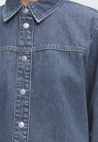 Μπλε τζιν μπουφάν με μεταλλικά κουμπιά, φαρδιά γιακά και οριζόντια ραφή στο στήθος.