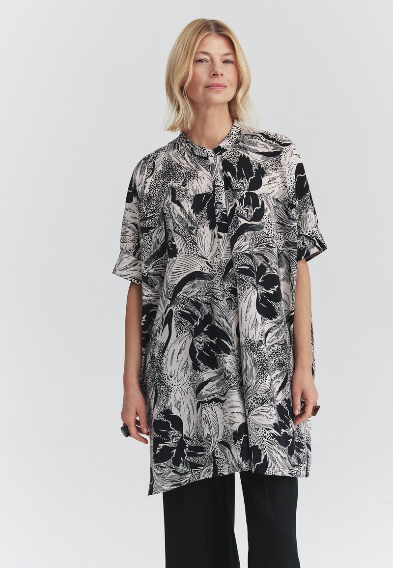 Femme aux cheveux blonds portant un chemisier ample à motif floral noir et blanc et un pantalon noir, debout devant un fond clair uni.