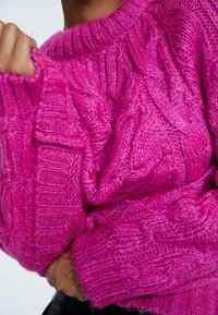 Fuchsia Kabelstrickpullover mit rundem Halsausschnitt und gerippten Bündchen. Der Stoff wirkt dick und strukturiert, mit ausgeprägten Kabelmustern.