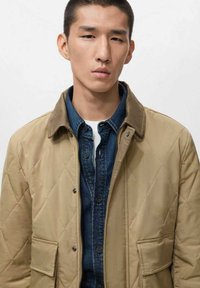 Giacca trapuntata di un beige chiaro con bottoni a pressione e tasche laterali, indossata sopra una camicia di denim scuro, con colletto e una maglietta bianca a girocollo sotto.