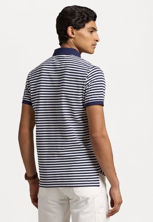 Striped polo shirt in navy and white, featuring a collar and short sleeves. Textured fabric with a fitted design, worn with light-colored pants.