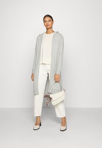 Grauer langer Cardigan mit Kapuze, cremefarbenes Oberteil, weiße Hose und beiger Handtasche mit gemustertem Band. Chunky Heels runden das Outfit ab.