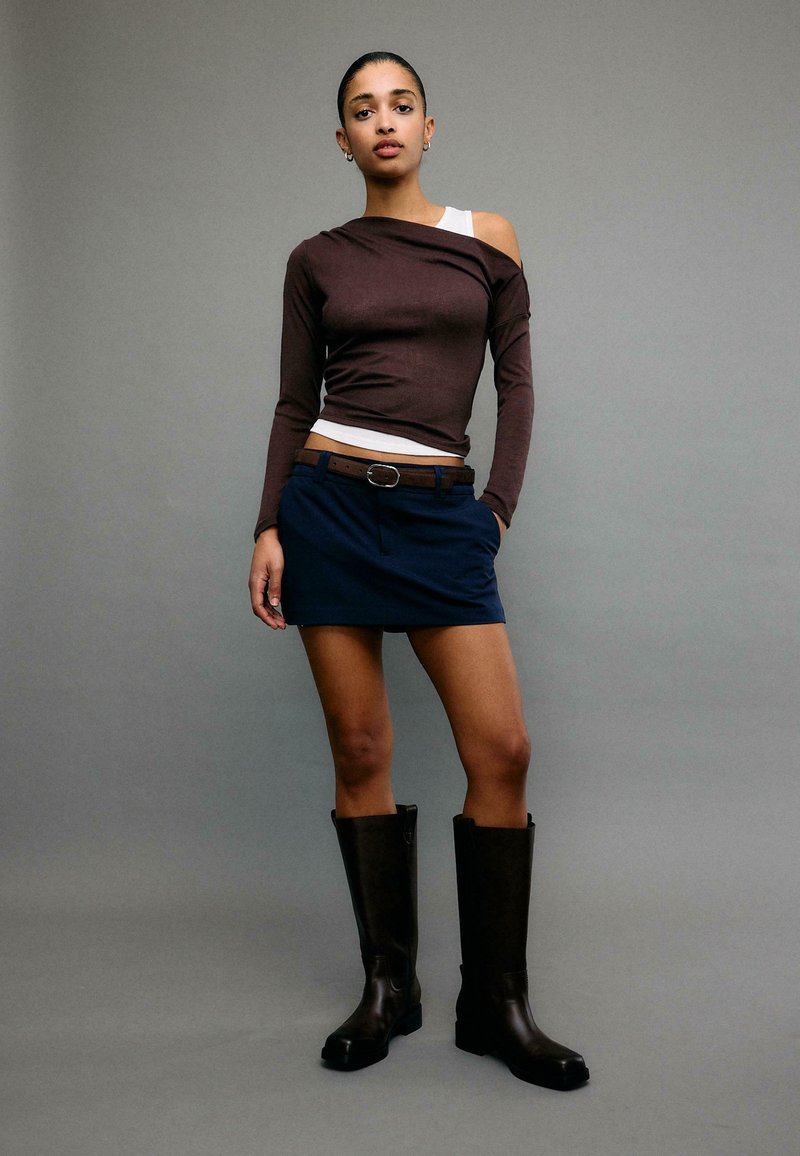 Femme debout devant un fond gris, portant un haut asymétrique marron, une mini-jupe marine avec ceinture et des bottes noires montant aux genoux.