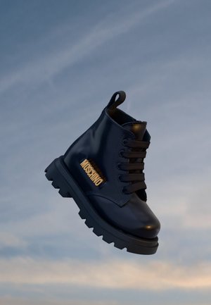 Czarny skórzany but bojowy z grubą podeszwą i czarnymi sznurowadłami, z złotym logo "MOSCHINO" po bokach na tle niebieskiego nieba.
