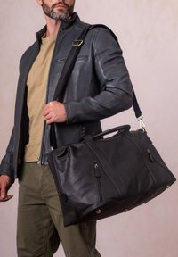 Black leather duffel bag with dual zippered pockets, top handles, and an adjustable strap, featuring a textured surface and gold hardware accents.