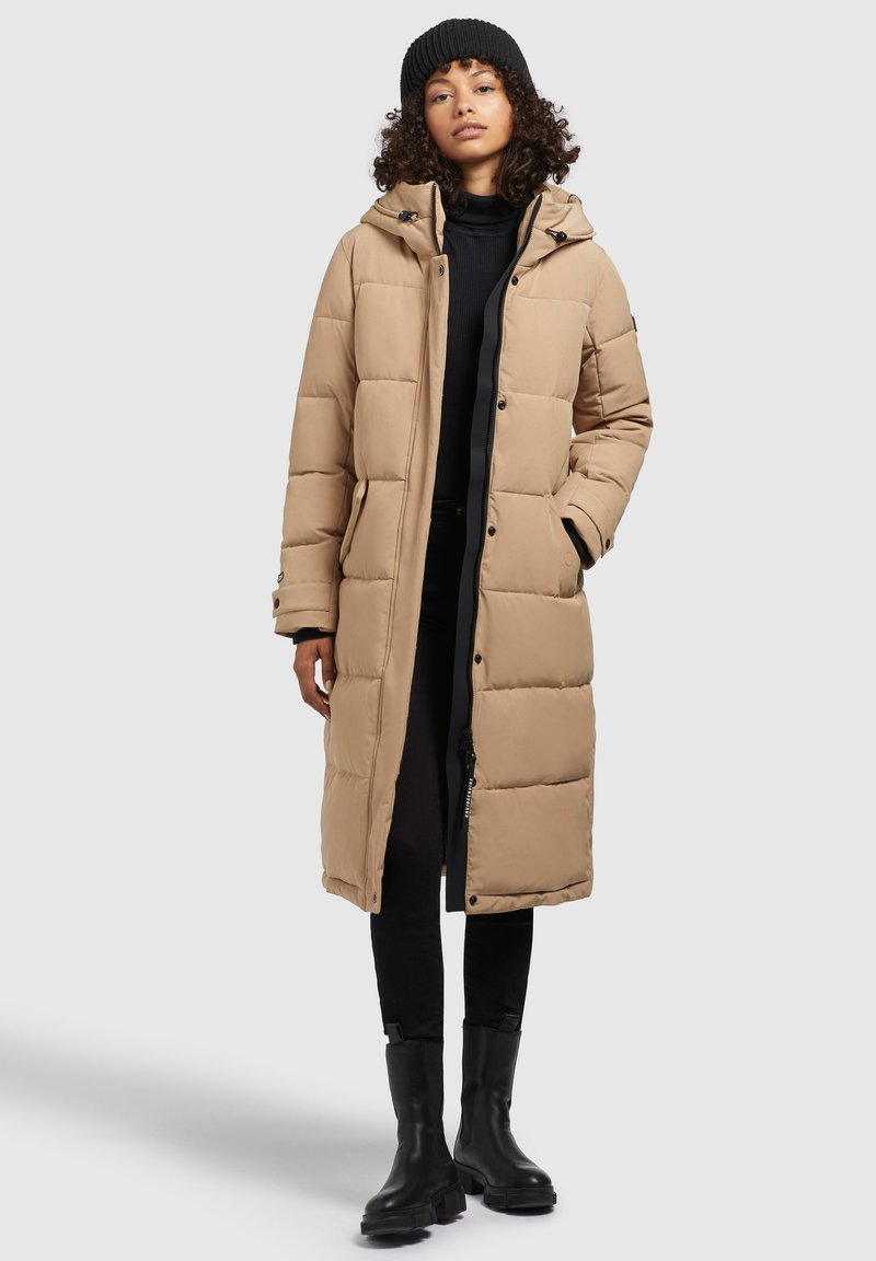 Longue doudoune beige avec capuche, dotée d'un design matelassé, fermeture par boutons et zip, et poches latérales. Portée sur une tenue noire.
