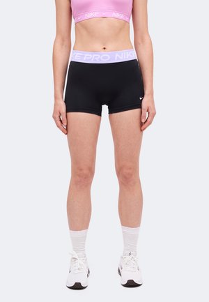 Un mannequin se tient devant un fond uni, vêtu d’une brassière de sport rose, d’un short Nike Pro noir, de chaussettes blanches et de baskets blanches.