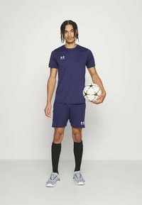 Mladý muž v námořnickém sportovním oblečení drží bílý fotbalový míč se vzory hvězd, stojí na jednobarevném světlém pozadí.