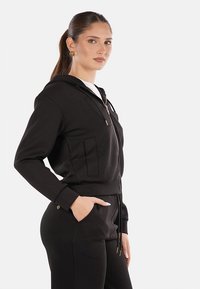 Felpa nera con zip, dotata di due tasche frontali, polsini a coste e fondo con coulisse. Realizzata in materiale morbido, con sottili dettagli in hardware dorato.