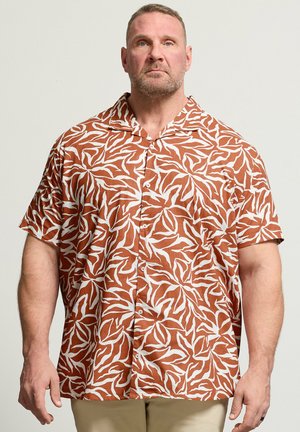 Homme aux cheveux courts et barbe portant une chemise à manches courtes à motifs de feuilles marron et blanche, debout devant un fond uni.