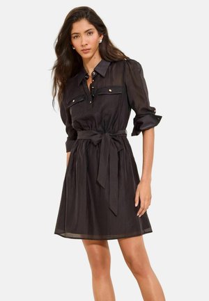 Frau mit langen dunklen Haaren, die ein schwarzes Hemdblusenkleid mit Gürtel und hochgekrempelten Ärmeln trägt, steht vor einem schlichten hellen Hintergrund.