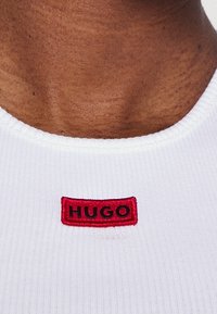 Närbild av en mörkhyad nacke som bär en vit ribbad tröja med en liten röd rektangulär patch med texten "HUGO" vid kragen.