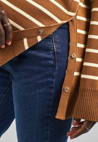 Maglione a righe marroni e crema, con bottoni laterali, abbinato a jeans blu scuro. Il maglione presenta una texture a coste e una vestibilità morbida.