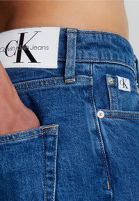 Siniset farkut, joissa on valkoinen nahkainen vyötärölabeli, jossa lukee "Calvin Klein Jeans" ja "ck"-logo, esittelevät näkyvää tikkausta ja taskudetaileja.