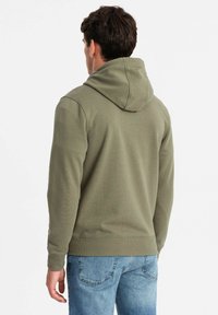 Sudadera color caqui con textura acanalada, capucha con cordón y bolsillo canguro; combinada con jeans de un azul claro.