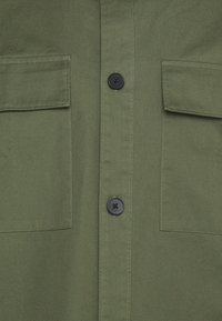 Oliwkowa koszula z dwoma kieszeniami na piersi, czarnymi guzikami i gładką teksturą, z zapinaniem na guziki z przodu oraz klasycznym kołnierzykiem.