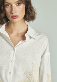 Camicia in lino bianco con colletto a ribadire, impreziosita da ricami floreali beige. Chiusura frontale con bottoni e vestibilità rilassata con maniche lunghe.