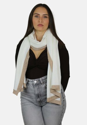 Femme aux longs cheveux noirs portant un haut noir, un jean taille haute gris clair et une écharpe blanche avec une bordure beige.