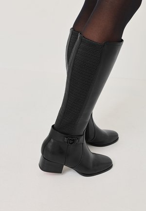 Black knee-high boots feature a textured mesh panel, smooth leather body, block heel, and buckle detail at the ankle.