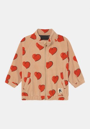 Veste beige zippée avec motifs de cœurs rouges, poignets et ourlet élastiques, poches avant et un petit patch logo chat noir en bas à gauche.
