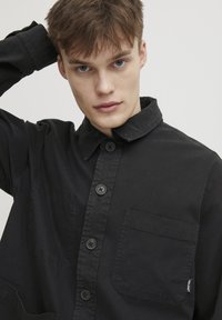 Black button-up shirt made of cotton with a collar, featuring a chest pocket and black buttons, smooth texture, and minimalist design.