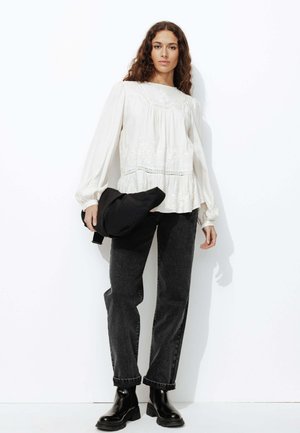 Blouse blanche brodée avec taille froncée et manches longues, associée à un jean noir taille haute et des bottines noires. Le mannequin tient un sac noir.