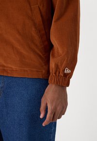 Brown corduroy jacket with elastic cuffs, featuring side pocket and logo detail. Worn over blue denim fabric, showcasing textured surface.