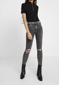 Mujer vestida con una blusa negra de manga corta con cremallera frontal, jeans ajustados grises rasgados y botas de tacón alto negras, de pie contra un fondo blanco.