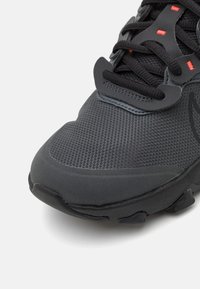 Scarpa sportiva nera con tomaia in rete testurizzata, suola in gomma e accenti rossi a contrasto nell'area dei lacci e nel marchio.