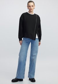 Zwarte sweatshirt met witte ronde hals en asymmetrische ritssluiting, gecombineerd met lichtblauwe wijde jeans en zwarte chunky laarzen.