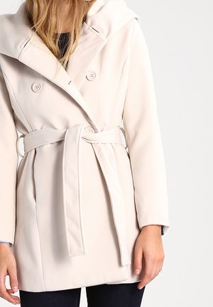 Vrouw draagt een lichtbeige mantel met ceintuur, grote kraag en dubbele knopenrij boven donkere broek, staand met ontspannen armen.
