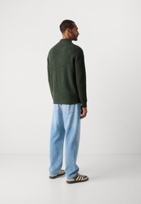Mann som står sidelengs iført en mørkegrønn strikkegenser, lyseblå vide bukser og grønne og hvite sneakers mot en ensfarget bakgrunn.